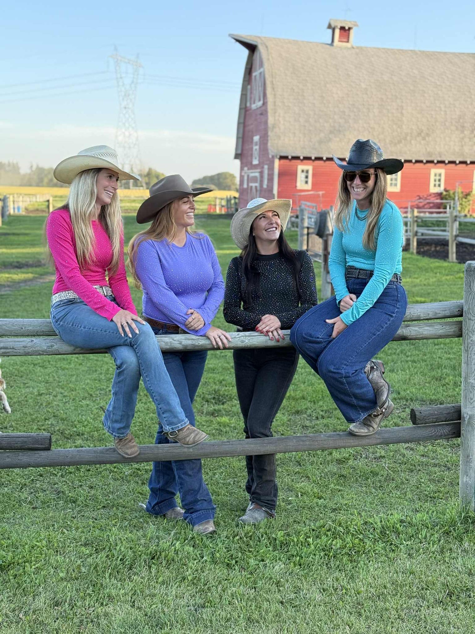 Four women wearing rhinestone bodysuits (pink, lavender, black, and turquoise) wearing cowboy hats sitting on a wooden fence at a barrel race.