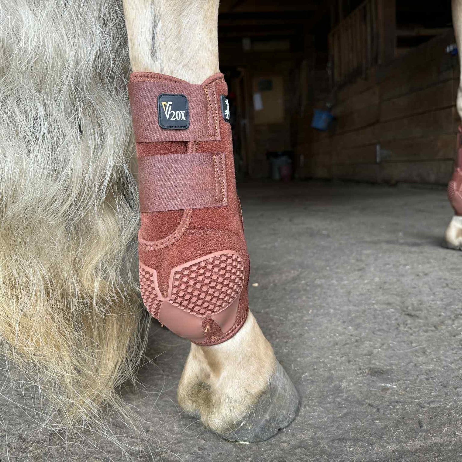 Horse's hoof with a brown protective boot labeled 'V20X' in a stable setting.