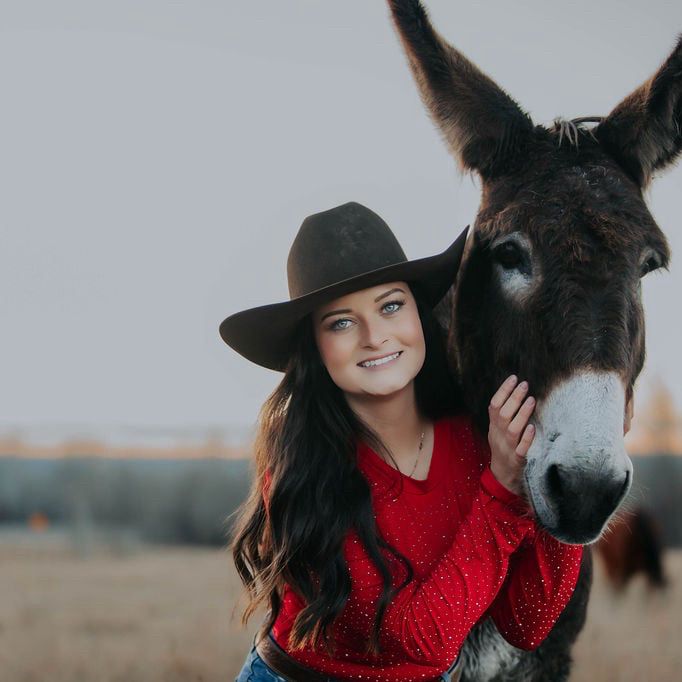 Woman in a red bodysuit and black hat petting a donkey outdoors.