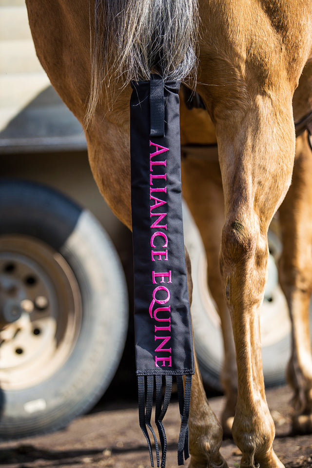 A black tail bag with the text 'Alliance Equine' written in pink, attached to a horse's tail.