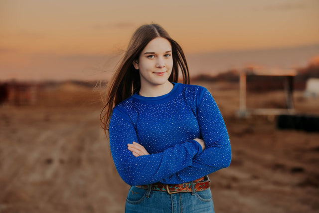 Woman in a Royal Blue rhinestone bodysuit and black cowboy hat standing in a field to match the Alliance Equine red V20x horse protective boots and bell boots