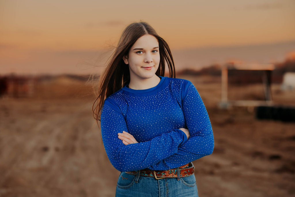 Woman in a Royal Blue rhinestone bodysuit and black cowboy hat standing in a field to match the Alliance Equine red V20x horse protective boots and bell boots