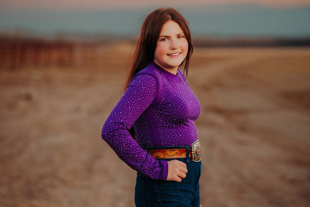 Woman wearing a purple long-sleeve rhinestone bodysuit to match her horses V20x protective horse boots and bell boots.