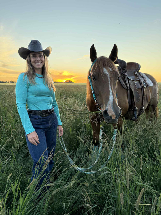 Woman in a cowboy hat wearing a turquoise bodysuit that matches her horse's turquoise Alliance Equine V20x hhorse boots and bell boots.