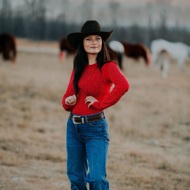 Woman in a red rhinestone bodysuit  and black cowboy hat standing in a field with horses in the background to match the Alliance Equine red V20x horse protective boots and bell boots