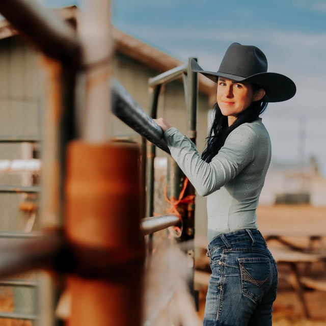 Woman in a cowboy hat and jeans standing, and Spruce Green bodysuit at a barrel race to match her Alliance Equine horses V20x boots and bell boots