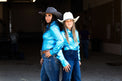 Two women posing in a dimly lit indoor space, wearing shiny Electric turquoise western-style jackets and jeans with cowboy hats, one in dark gray and one in white.