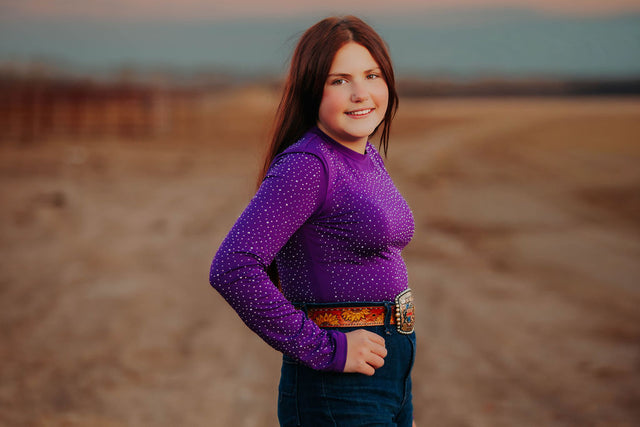 Woman wearing a purple long-sleeve rhinestone bodysuit to match her horses V20x protective horse boots and bell boots.