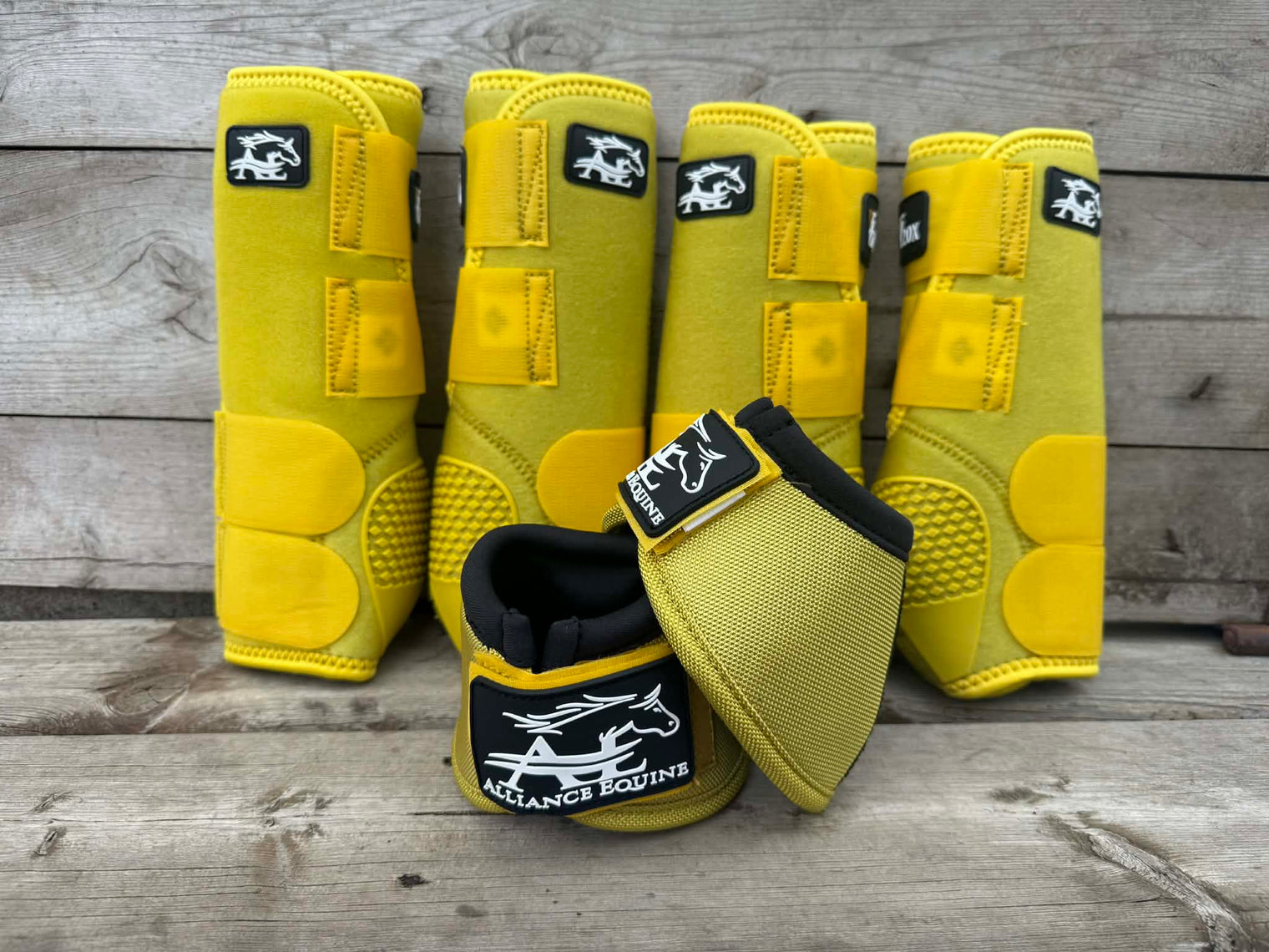 Four yellow horse leg boots and one matching horse boot wrap arranged on a rustic gray wooden plank surface. The boots feature black and white branding patches for 'Alliance Equine'.