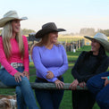 Four women in cowboy hats sitting on a wooden fence with a rural background wearing Rhinestone bodysuits